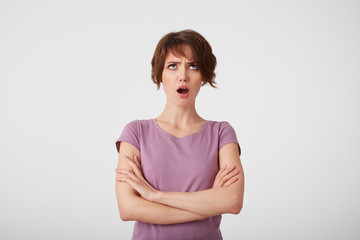 Obraz premium Portrait of frowning short-haired lady in blank t-shirt, offended looks up, looks sad and unhappy, stands over white background with crossed arms.