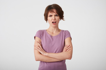 Portrait of frowning displeased short-haired lady in blank t-shirt, outraged by too low exam grade, stands over white wall with arms crossed.