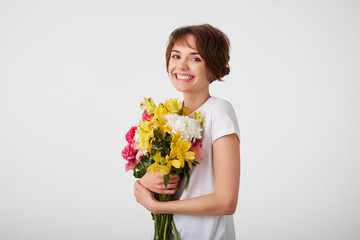 Portrait of young smiling cute short haired girl in white blank t-shirt, holding a bouquet of colorful flowers, very glad to such a gift from her boyfriend, broadly smiling over white wall.