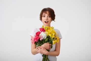 Portrait of happy amazed young cute short haired girl in white blank t-shirt, with wide open mouth and eyes, holding a bouquet of colorful flowers, surprised by such a gift, isolated over white wall.
