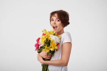 Young happy amazed cute short haired girl in white blank t-shirt, with wide open mouth and eyes, holding a bouquet of colorful flowers, surprised looking at the camera isolated over white wall.