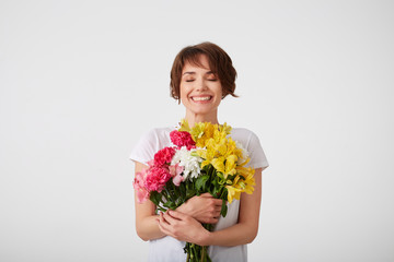 Happy cute young short haired girl in white blank t-shirt, holding a bouquet of colorful flowers with closed eyes, smiling broadly, standing over white background.