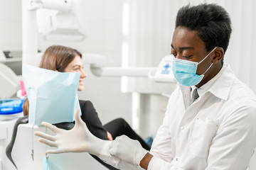 The doctor puts on rubber gloves. Young African male dentist with a patient. Woman in the dentist chair at dental clinic. Medicine, health, stomatology concept. dentist treating a patient.