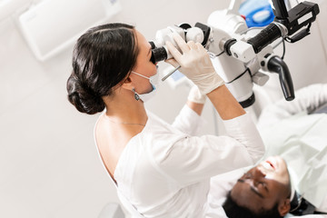 Young woman dentist treating root canals using microscope in the dental clinic. Man patient lying...