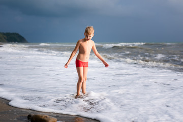 Child boy playing in the waves on the beach in summer sunset, kid watching sea waves and having fun
