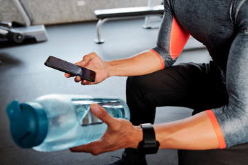 Close-up image of sportsman checking social media on smartphone and drinking water during break in gym