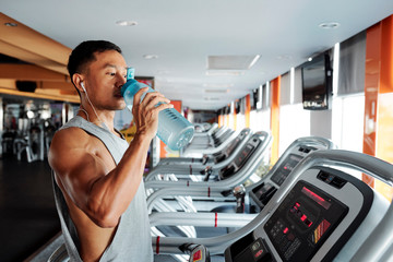 Muscular Asian sportsman drinking water after running on treadmill in gym