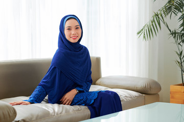 Pretty young Muslim woman in blue press and hijab resting on sofa at home