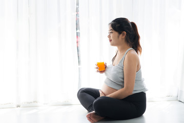 Asian pregnant woman holding a glass of orange juice and sitting near window, lifestyle concept.