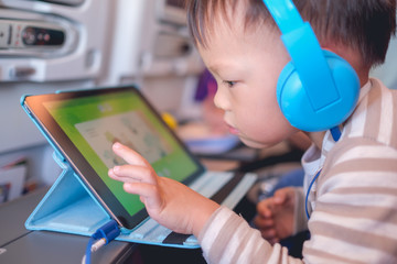 Cute Asian 2 - 3 years old toddler boy child wearing headphones using tablet pc watching cartoons / playing game during flight on airplane. Happy Flying with children concept, soft & selective focus