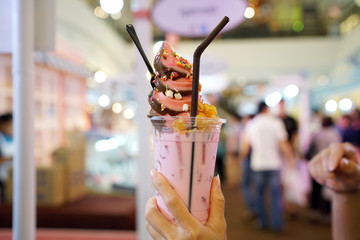 Holding a plastic glass of pink fresh milk topped with two tone soft serve ice cream, Charcoal and strawberry flavors.