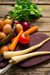 basket of eggs and tomato, spanish onions, carrots, parsley roots, celery, cutting board, old weathered wooden table background