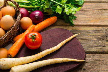 basket of eggs and tomato, spanish onions, carrots, parsley roots, celery, cutting board, old weathered wooden table background