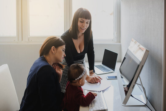 close up photo of two women in office outfit working on computers and discussing a report on the floor; one of them is with a baby girl in her arms