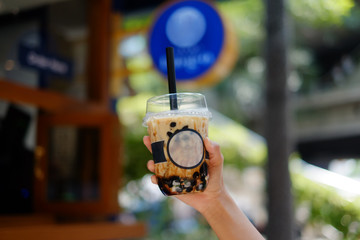 Hand Holding a plastic glass of refreshing Milk Tea Latte with bubble (boba), selective focus. blue...