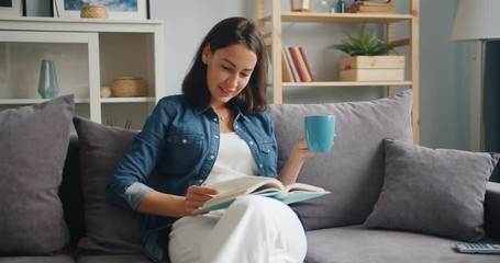 Slow motion of attractive girl reading book holding cup of coffee at home relaxing on comfortable sofa. Youth lifestyle, people and apartment concept.