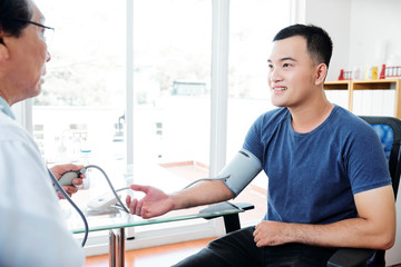 Doctor measuring blood pressure of young smiling Asian man