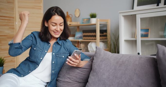 Excited young lady celebrating success raising arm looking at smartphone screen smiling at home getting great news. Happy people, gadgets and youth concept.