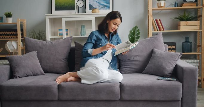 Slow motion of beautiful girl in casual clothing reading book relaxing at home sitting on couch in nice apartment. Youth lifestyle, house and people concept.