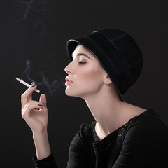 Obraz premium Young fashion woman smoking cigarette in hat and black drees over dark background. Vintage female portrait, styling. Image toned.