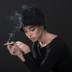 Obraz premium Young fashion woman smoking cigarette in hat and black drees over dark background. Vintage female portrait, styling. Image toned.