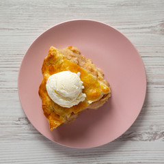 Slice of homemade apple pie with ice cream on a pink plate over white wooden surface, overhead view.