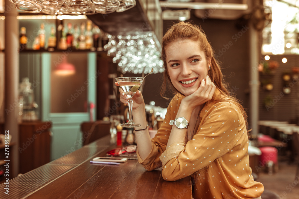 Wall mural smiling woman holding martini drink sitting at the bar counter