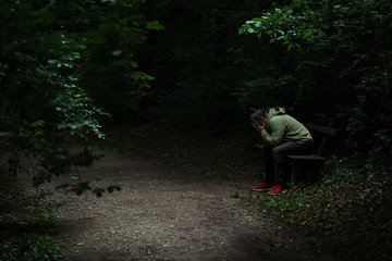 Man sitting on a bench in light spot on forest path, surrounded with darkness, covering his face with his hands, alone, sad and depressed