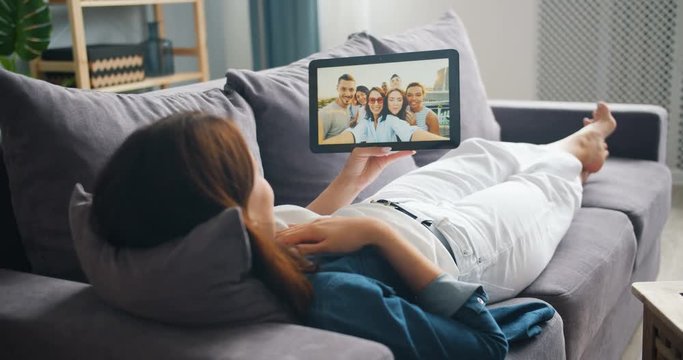 Young woman is making online video call with tablet talking to friends lying on couch at home enjoying communication. People, gadgets and apartment concept.