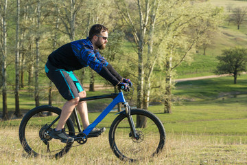 Cyclist in shorts and jersey on a modern carbon hardtail bike with an air suspension fork rides off-road on green hills near the forest	