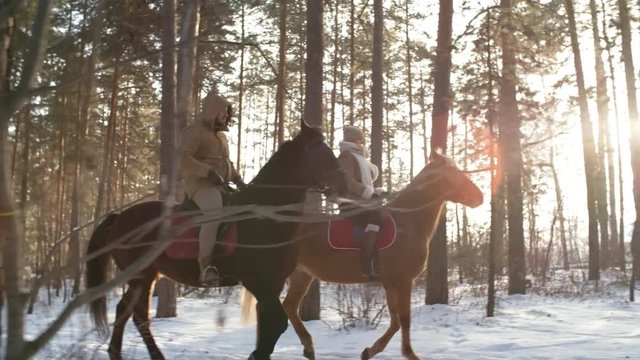 Side view of man and woman riding horses in pine forest on sunny winter day