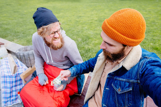 Sneaky, sly, liar man plotting something helping beggar making selfie with homeless while presenting coints in city street. They sits together on bench. Human emotions, facial expressions, attitude