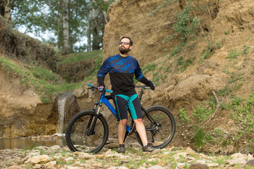Fototapeta premium Cyclist in shorts and jersey on a modern carbon hardtail bike with an air suspension fork stands on the rocky shore of a mountain river 