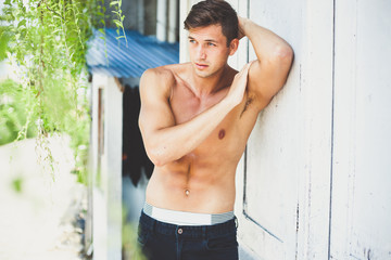 Fototapeta na wymiar Portrait of a beautiful sexy brunette with nude sport torso in black jeans. a young man stands outside leaning on a white wall. relief biceps and press. sunny day