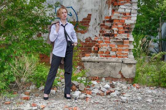 Woman of androgynous appearance and short blonde hair bob hairstyle with coat in hand, men's white shirt, tie and trousers, smoking a cigarette with an abandoned ruins on background. Noir detective.