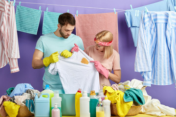 housewife and husband looking at spot T-shirt while standing behind table with clothes and bowl. service concept.isolated blue background. man and woman thinking about how wash dirty clothes
