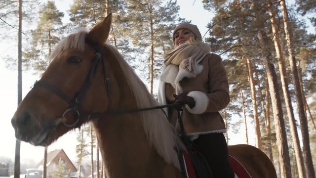Low angled shot of happy caucasian woman sitting on horse, looking at camera and smiling in forest on winter day