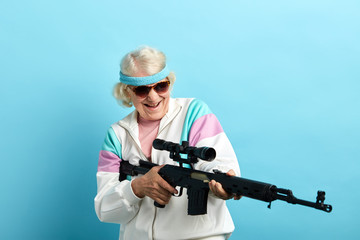 Senior woman in sports wear and black sanglasses screaming while aiming at camera with a rifle isolated on blue.