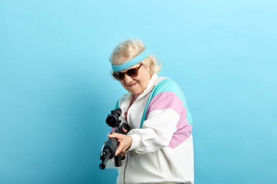 Senior woman in sports wear and black sanglasses screaming while aiming at camera with a rifle isolated on blue.