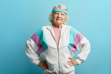 Optimistic elder female coach or trainer dressed in light sports suit and headband posing in studio on blue background, copyspace