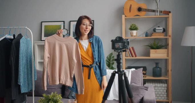 Joyful young woman blogger recording video about clothes talking gesturing at home using professional camera on tripod. Youth, occupation and technology concept.