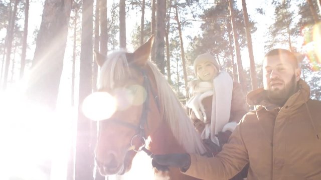 Low angled shot of woman riding horse and speaking with man while he holding leading reins and walking in forest on sunny winter day; sunlight shining at camera