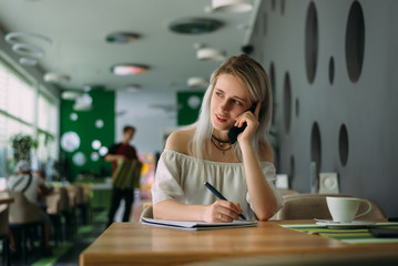 nice-looking woman sits in a restaurant and makes a business call and notes in notepad.
