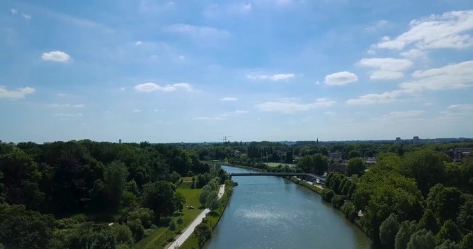 Aerial drone vertical flight over a canal and a wooded park that lets discover the city in the background. 4K