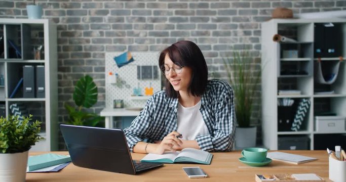 Attractive young woman is chatting in skype using laptop in office holding cup of tea enjoying conversation and coffee break. People, communication and job concept.