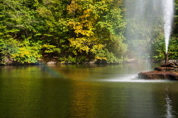Fototapeta na wymiar Water jet fountain on rock in center of lake with colorful fall trees reflected in calm water surface. Rainbow in water flow. Landscape of Sofiyivka park in Uman Ukraine. Autumn in Sofievka Uman park 