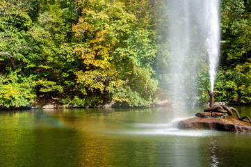 Fototapeta na wymiar Bronze snake fountain on rock in center of lake with colorful fall trees. Rainbow in water drops from fountain flow. Landscape of Sofiyivsky Park in Uman Ukraine. Autumn in Sofievka Uman park