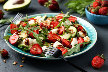 Healthy salad with strawberries, avocado, arugula and mozzarella, dressed with olive oil and balsamic dressing located on a dark background
