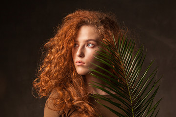 Energizer beautiful young curly ginger hair woman with green tropical plant palm branch posed in dark walls studio. Copy space background.