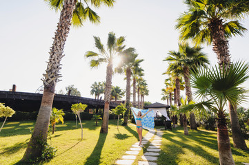 Fototapeta premium Beautiful happy young slim girl stands near Tall palm trees. In the blue dress, shawl pareo. Turkey. Palms. recreation. Vacation. Sunlight.